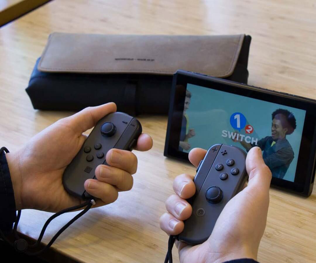 Hands holding Joy-Con controllers next to Nintendo Switch and brown console case