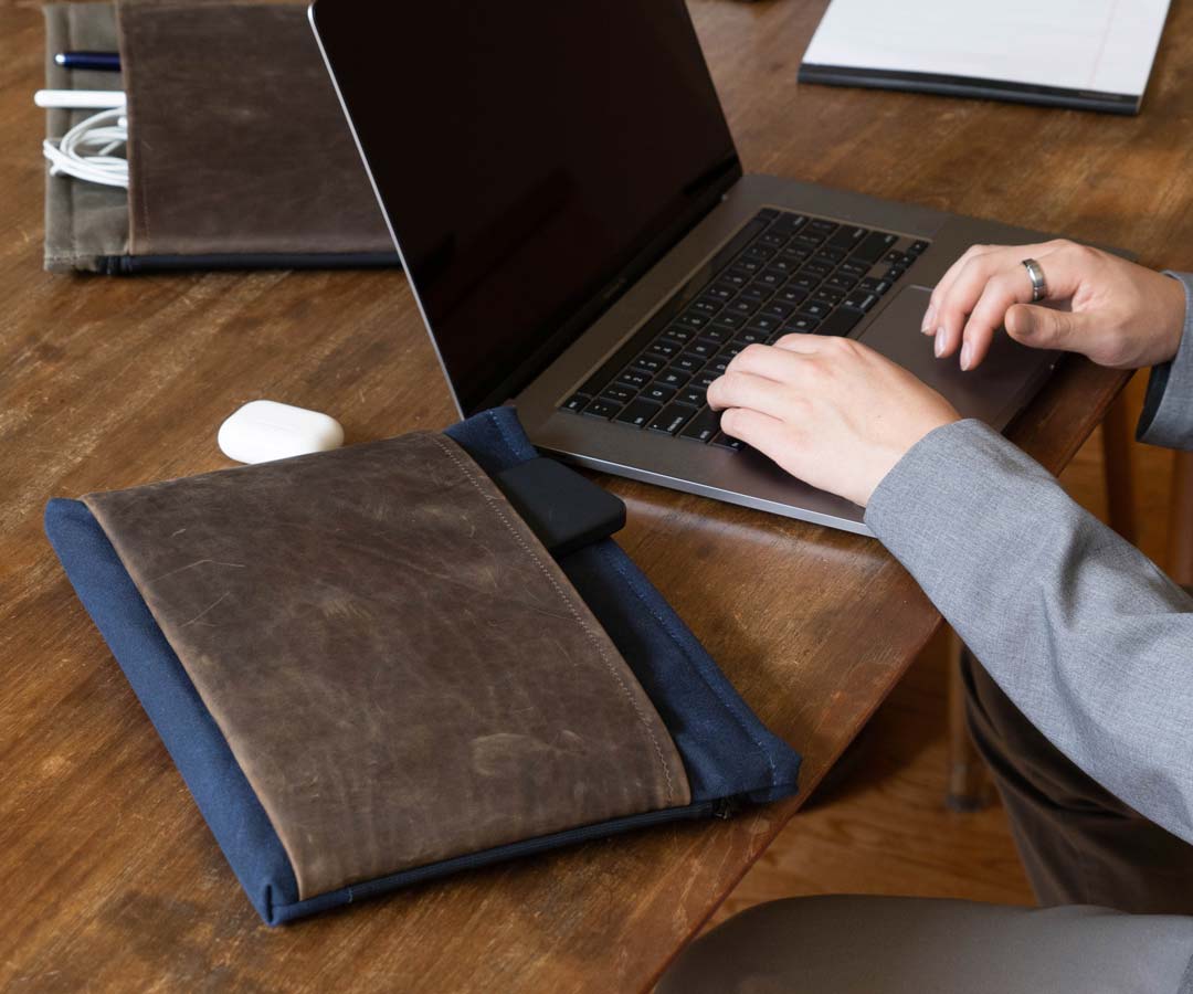 Person placing MacBook into brown leather sleeve on wooden desk