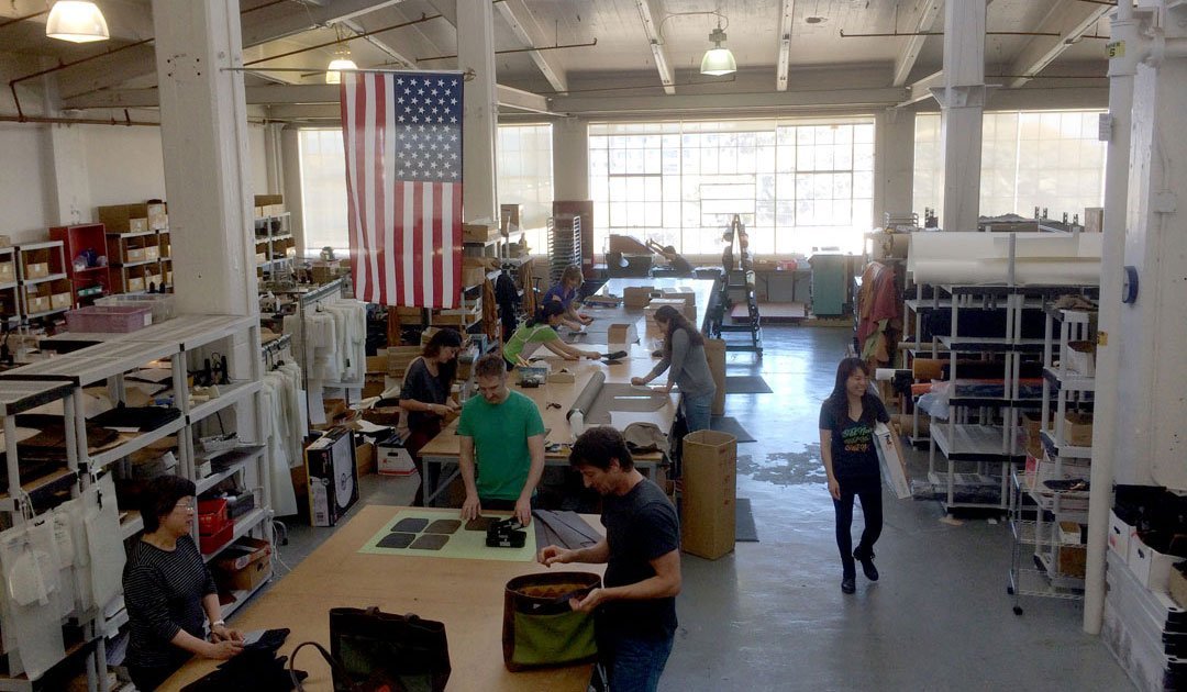 Artisan workshop with team members assembling bags under a large American flag in a sunlit factory space