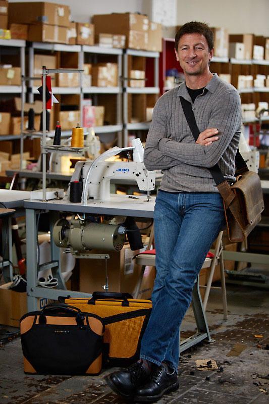 Gary Waterfield smiling in his workshop, leaning on a sewing machine table surrounded by shelves, boxes, and leather bags